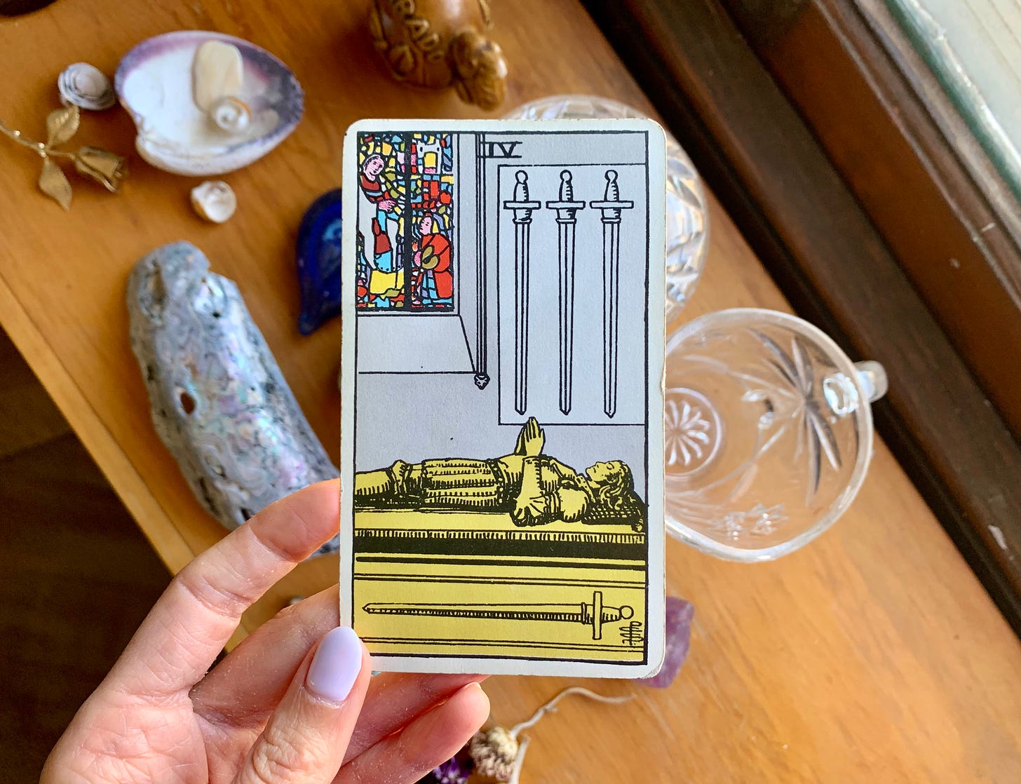 A hand is holding a tarot card, four of swords by pamela colman smith. In the image, a person is laying in a sarcophagus with their hands folded in prayer position over their chest. There is a stained glass window and three swords on the wall. Behind the card is a wooden shelf with various items on it, some shells, a gold rose pin, a vintage sugar bowl. 