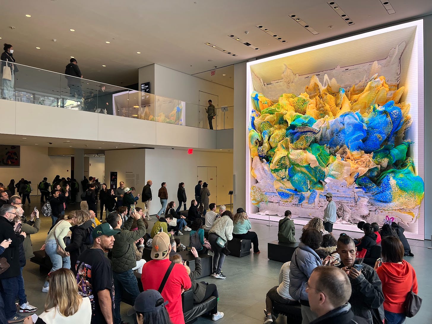 A crowd of visitors gathers in front of Refik Anadol's piece "Unsupervised" at the Museum of Modern Art. The piece has green, yellow, and blue pulsating blobs, and is two stories tall