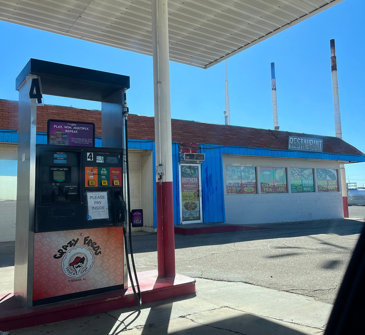 A gas station. The logo under a gas pump reads "Crazy Fred's" and has a cartoon man with a funny face on it. Behind is a building that says "BROTHERS" on the door. There's a sign that says RESTAURANT, and a bunch of window stickers with the specials: chicken curry, roti, mango lassi.