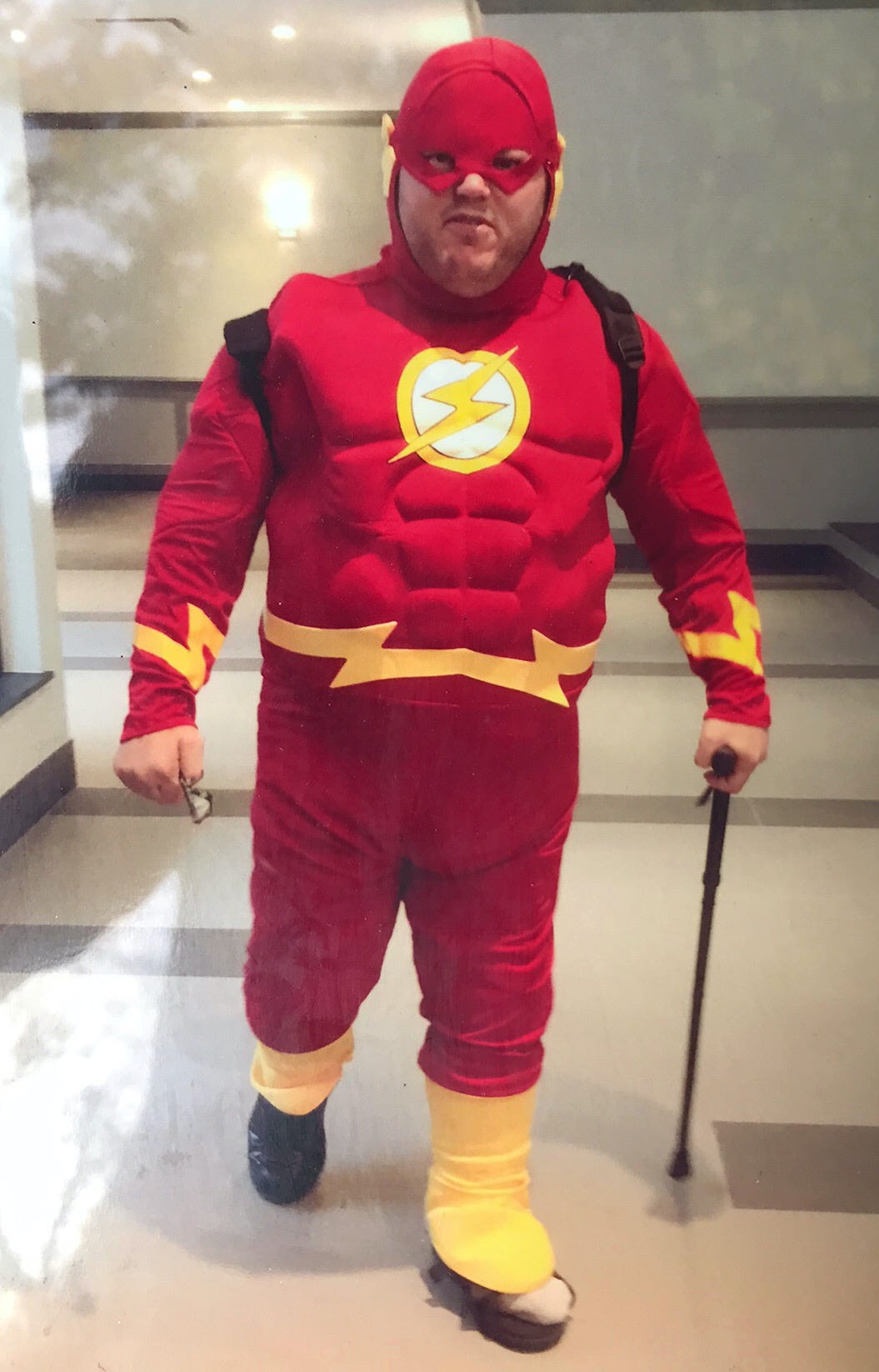 A smiling man wears a red-and-yellow costume to portray the superhero The Flash. The yellow logo on his chest looks like a lightning bolt. He stands in a building lobby, and is using a cane. On his shoulders we can see the straps of the knapsack he is wearing. He has taken off his glasses, which he holds in his right hand.