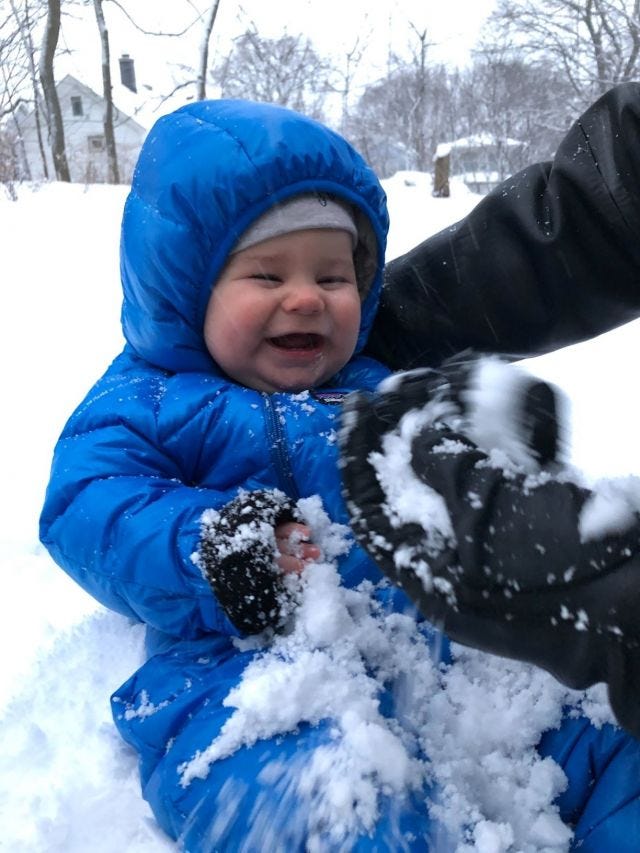 a chubby-cheeked infant in a snowsuit laughing at snow