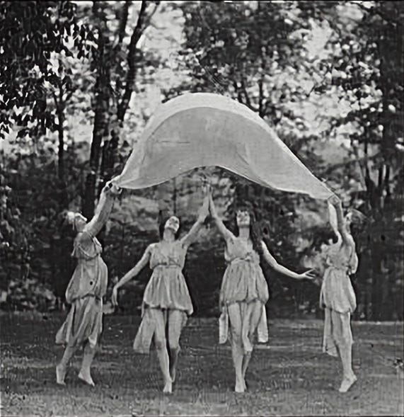 a 1915 black and white photo shows four female dancers in tunics throwing up a large piece of fabric in the air in an outside setting
a 1915 black and white photo shows four female dancers in tunics throwing up a large piece of fabric in the air in an outside setting