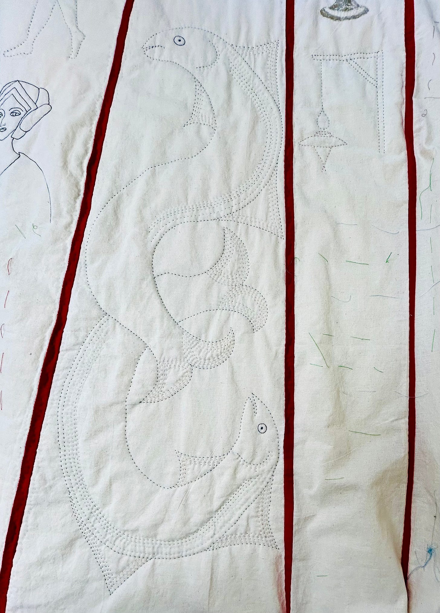 Two large, long fish, quilted onto cream fabric. The fish are intertwined. Other motifs, including a woman’s head can be seen to the side of the fish, but the fish are the main focus of the image. 