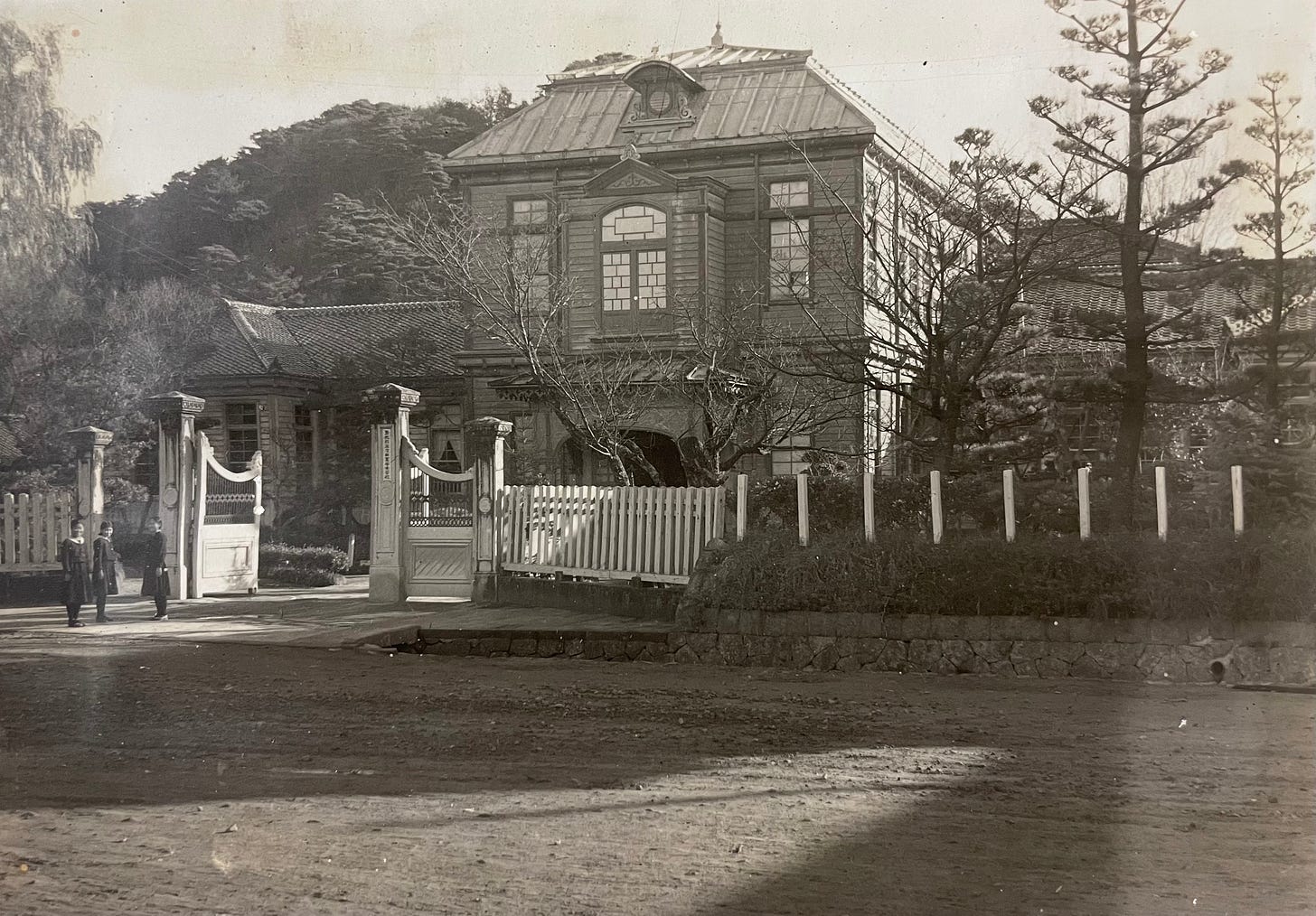 a black and white photo of a large, old two story building behind a large gate. Three girls are standing in front of the open gate