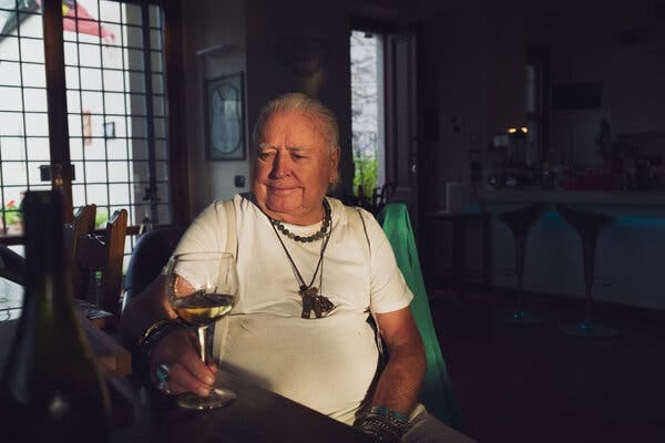 A man with white hair and a number of necklaces around his neck holds a glass of wine at a table in his home.