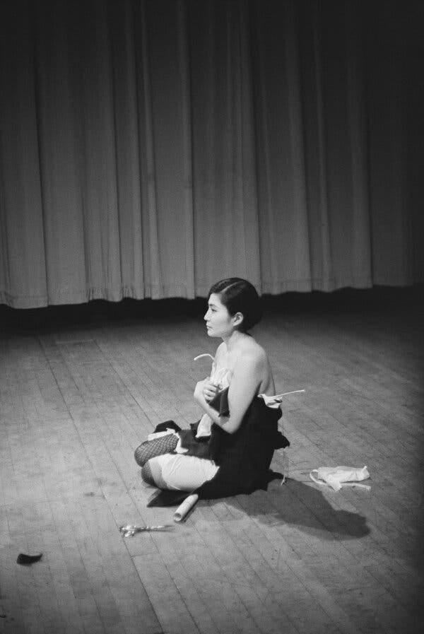 A black-and-white photo shows Yoko Ono sitting on a wooden floor with a curtain behind. Her clothes have been cut away so that her arms are bare, and there are fabric scraps on the floor around her.