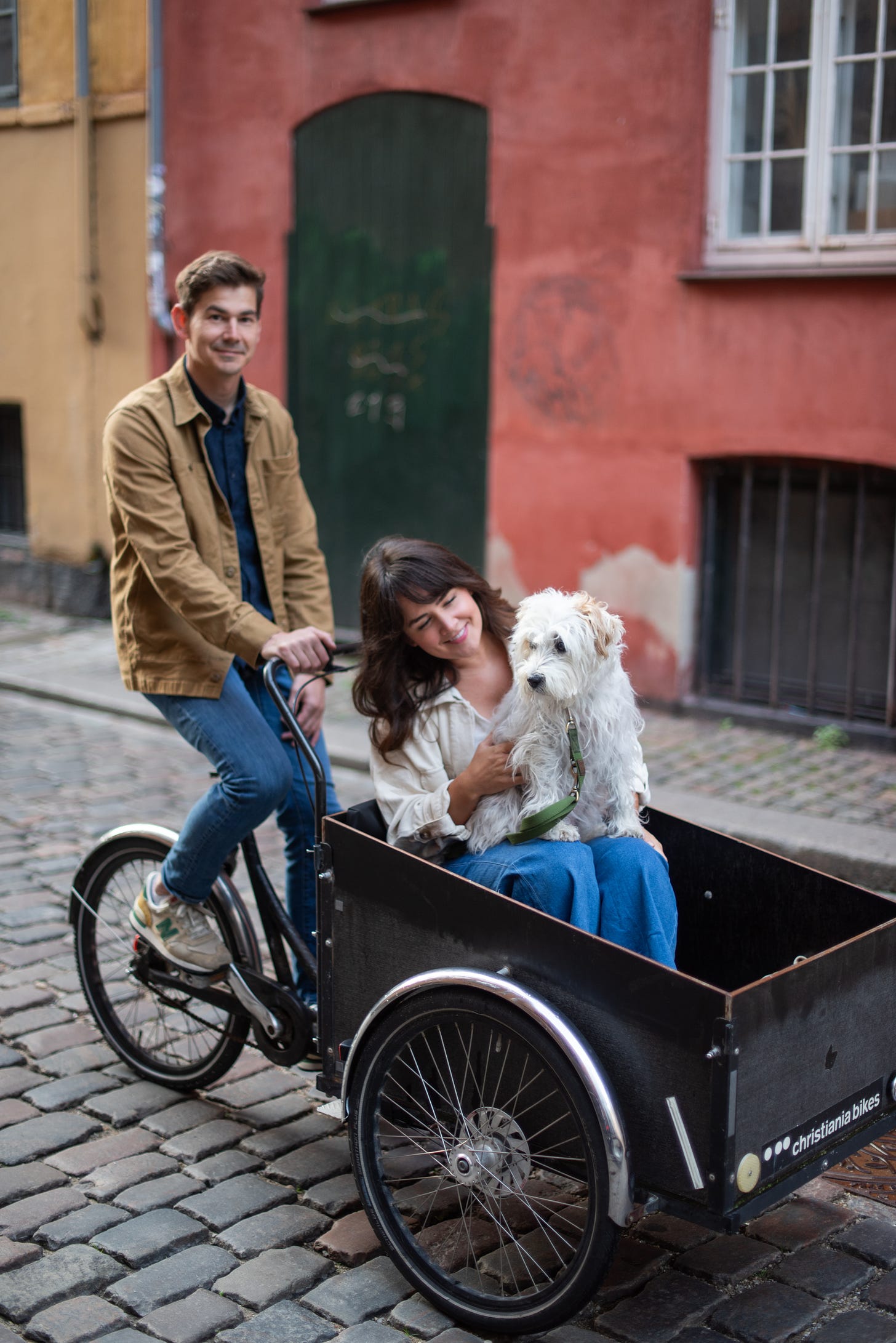 The author, her partner, and their dog riding down a cobblestone street in a cargo bike. The are in front of a red building and are smiling at the camera.