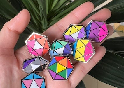 a handful of D20-shaped enamel pins in pride flag colors