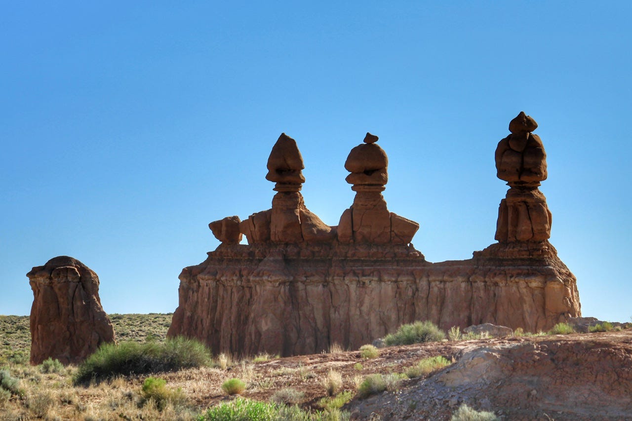 A red rock monument with three hoodoos stands above a desert floor with a clear blue sky as a backdrop.