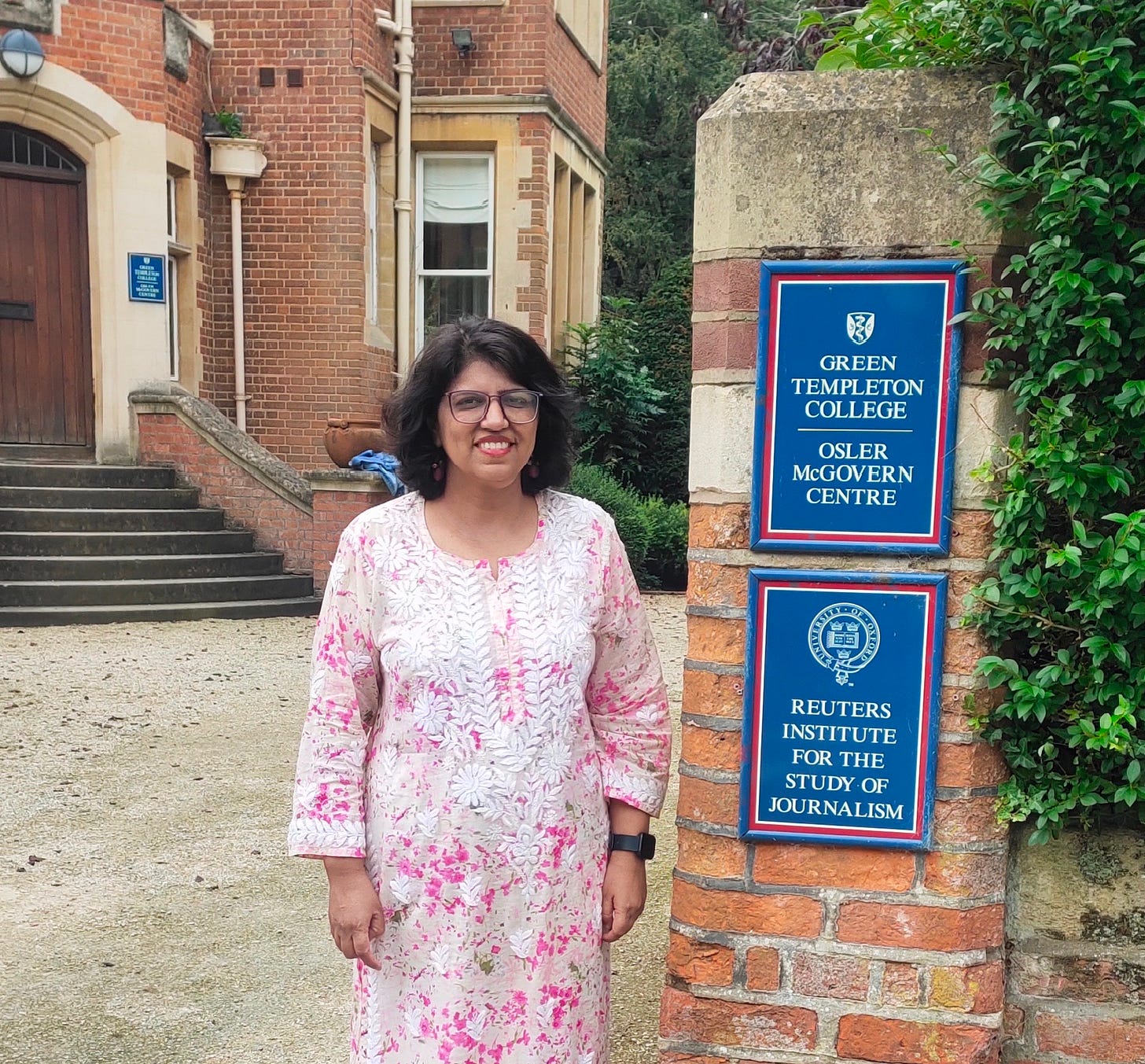 That’s me, Priti, an Indian woman standing outside the Reuters Institute for the Study of Journalism in Oxford. My pink floral kurta perfectly captures the essence of a rare bright day in Oxford. The pillar on my left has two boards, one with the name of the Institute and the other with Green Templeton College and Osler McGovern Centre. Behind me is the Institute’s building and visible prominently are the steps to the entrance door. I’m, as usual, wearing my pink-rimmed glasses and smiling.