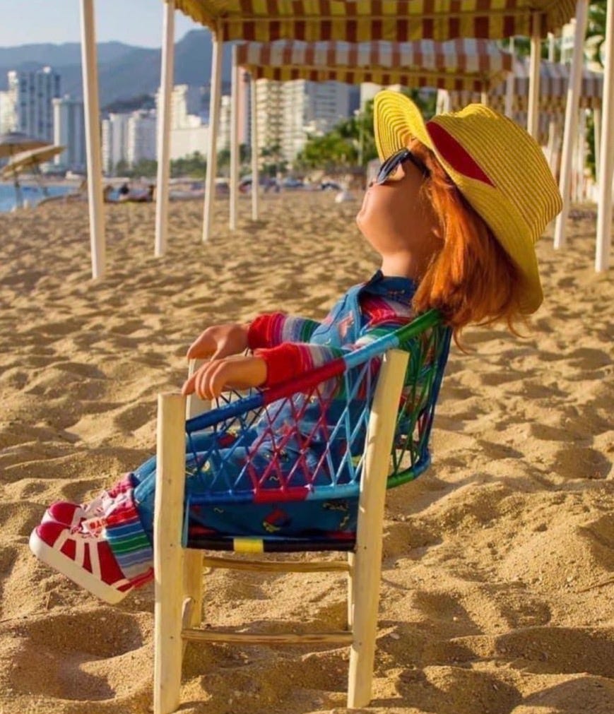 chucky doll in a chair on a beach with glasses and straw hat on, looking relaxed