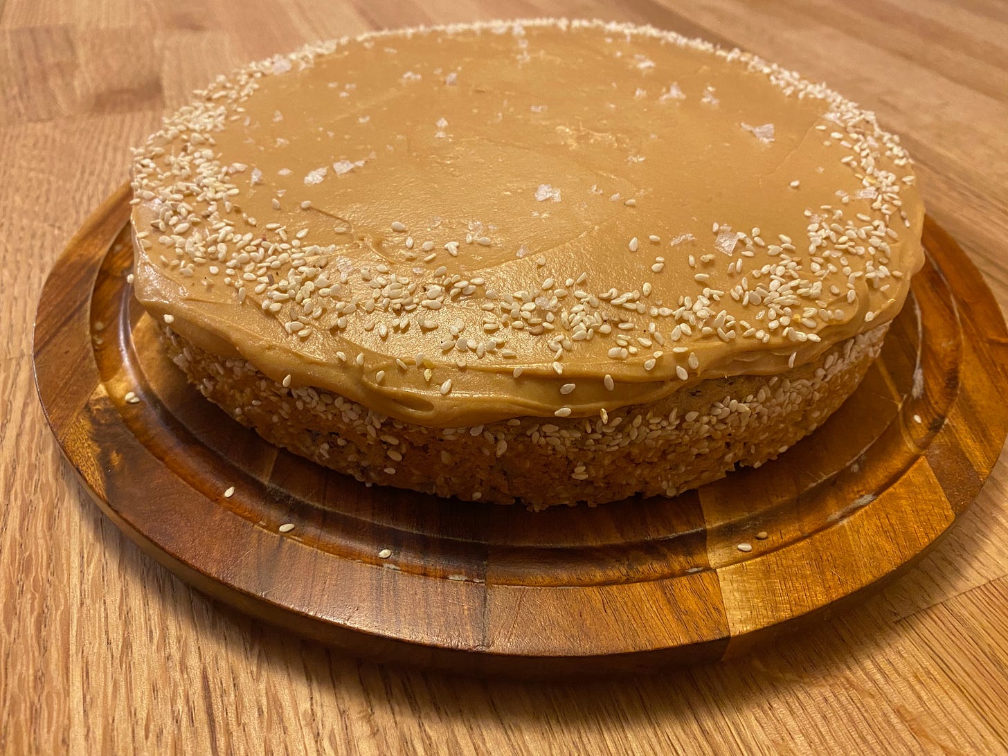 A round cake with its sides covered in sesame seeds sits on a wooden platter. The top is coasted in a thick frosting and sprinkled with sesame seeds.