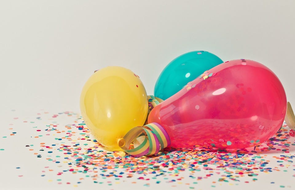 Foto de três balões sobre uma superfície branca repleta de confetes coloridos de papel. Da esquerda para a direita, os balões são amarelo, azul e vermelho, e no centro dos três, aparecendo em primeiro plano entre o amarelo e o vermelho, há uma fita com as cores verde amarelo e rosa, enrolada em si mesma. O fundo da imagem é branco.