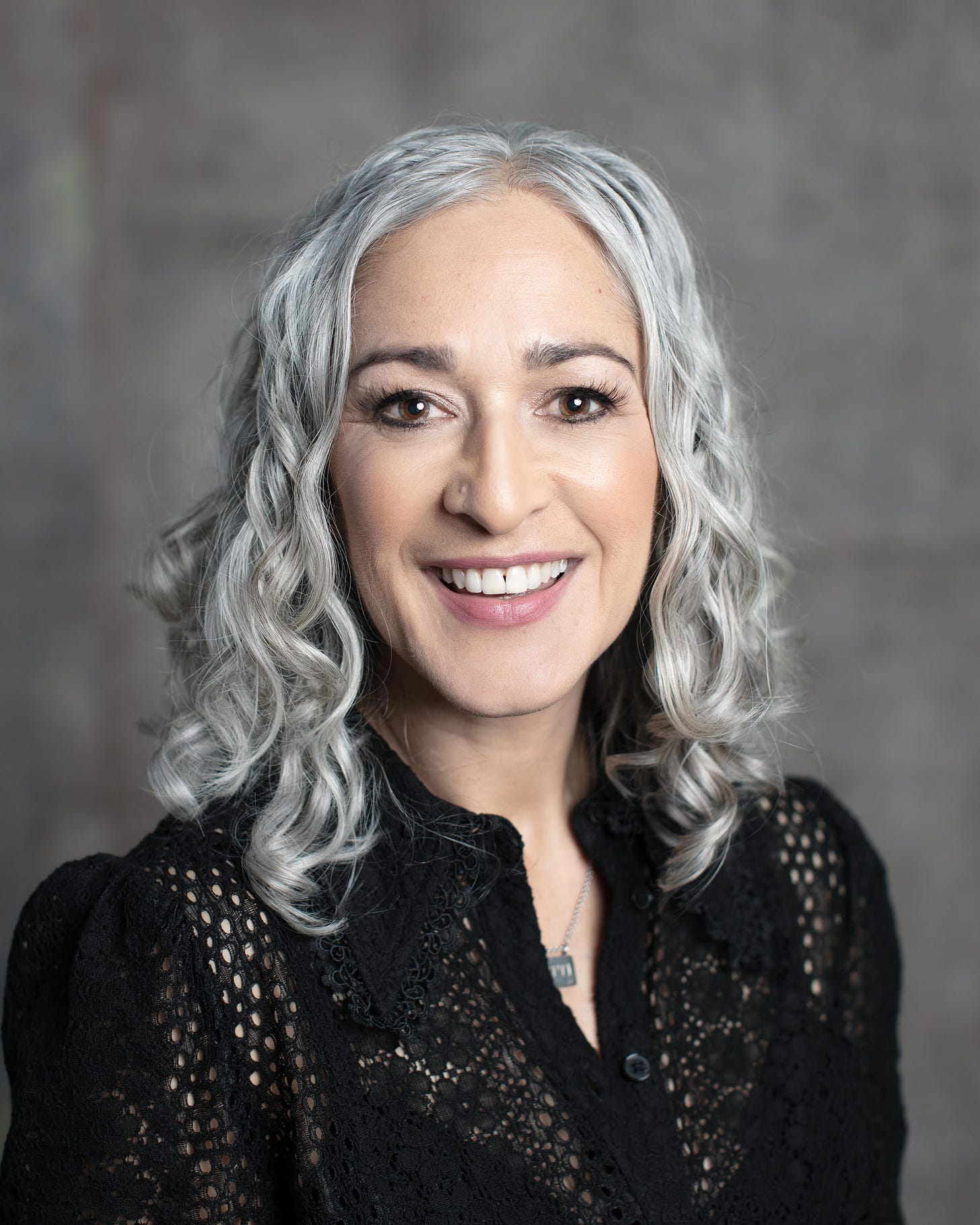 A person with light skin and medium-long, wavy light-grey hair smiles at the camera while wearing a flower-latticed black top, set in front of a blurry, textured grey background.