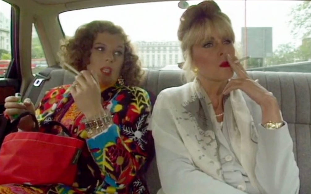 Patsy and Edina sit in the backseat of a large car both smoking cigarettes