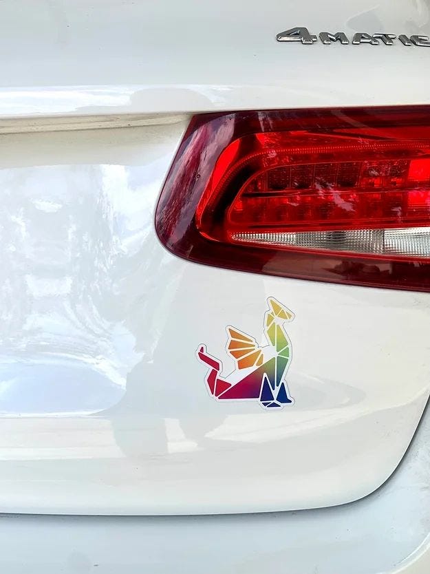 A small rainbow dragon made up of geometric shapes is shown on the back hatch of a white car. A small rainbow dragon made up of geometric shapes is shown on the back hatch of a white car.