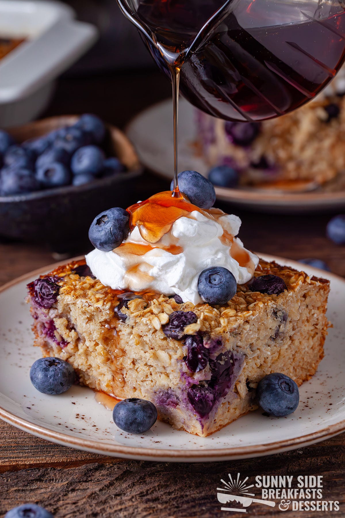 Pouring maple syrup onto a sliced of baked blueberry oatmeal with whipped cream.