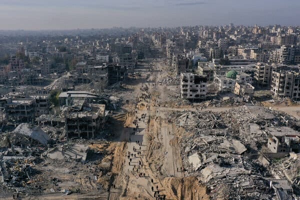 An aerial view shows a stream of people walking into a city in ruins. 