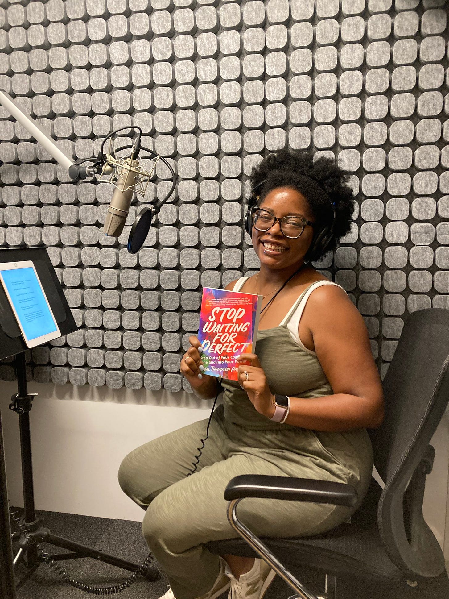 Black woman in a recording booth wearing headphones and dressed in green overalls holding a book