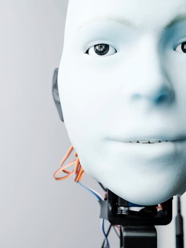 A humanlike robot face, made of a light blue material and with and a slightly open mouth, gazes directly at the reader. Its eyes are small cameras, and wires extend from where the neck would be.
