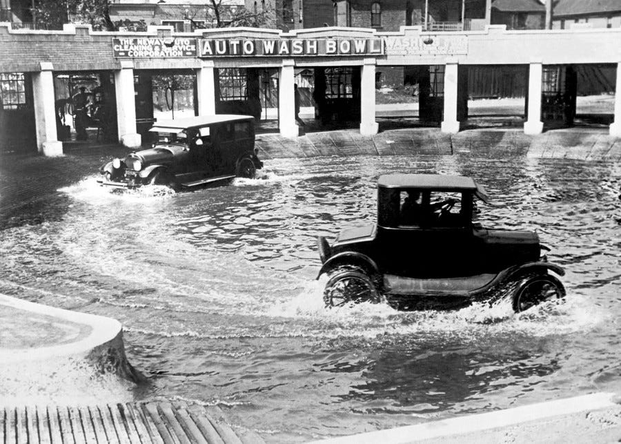 Two older passenger cars drive through water in a shallow pool to clean their underside.