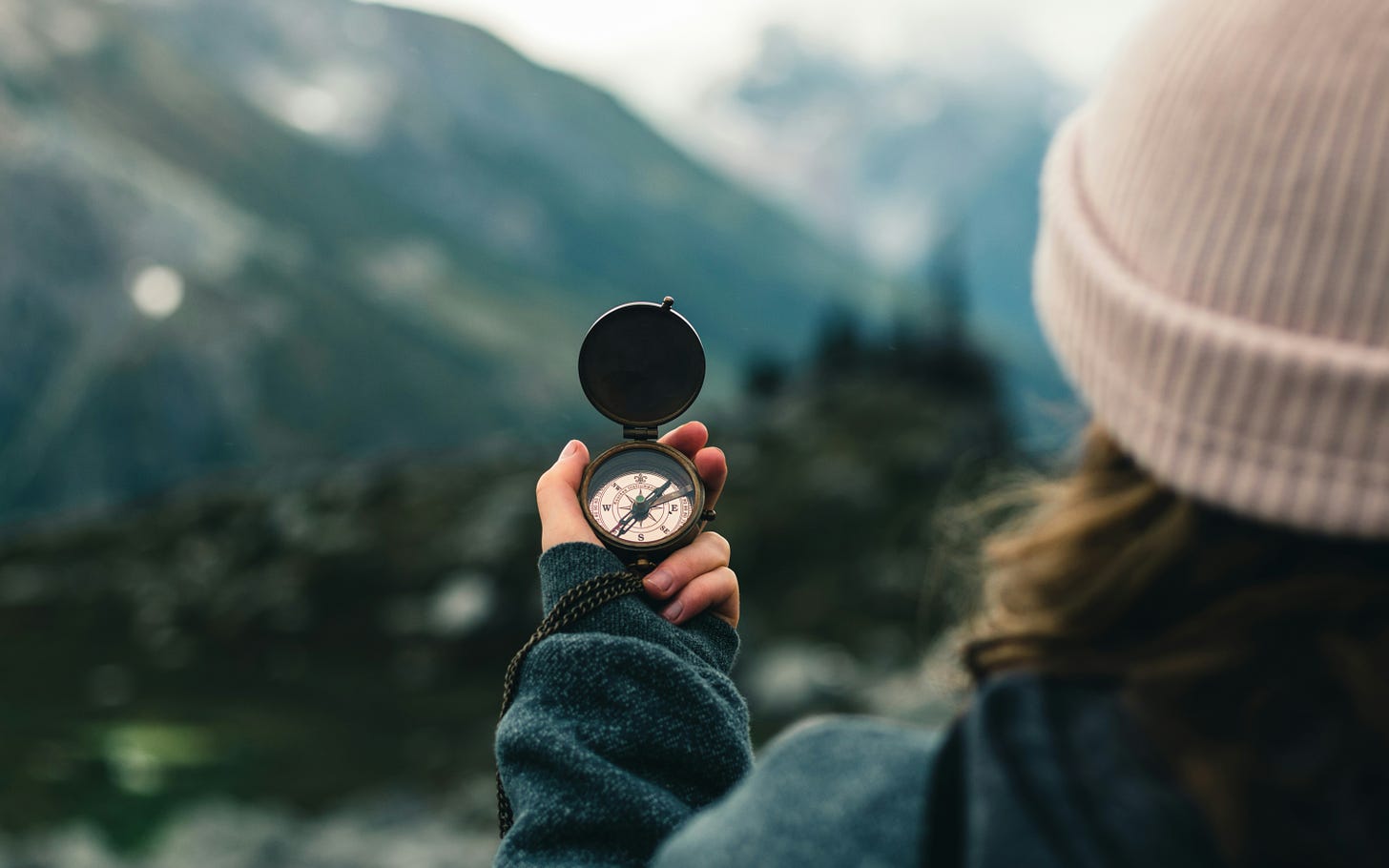 A person holds a compass in their out stretched arm. In front of them, out of focus, are mountains and trees.