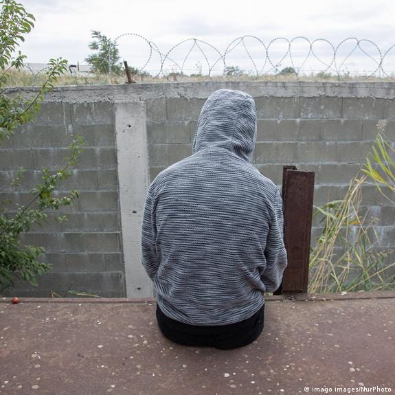 A boy wearing a grey hoodie, with his back turned to the photo, sits before a high wall with barbed wires
