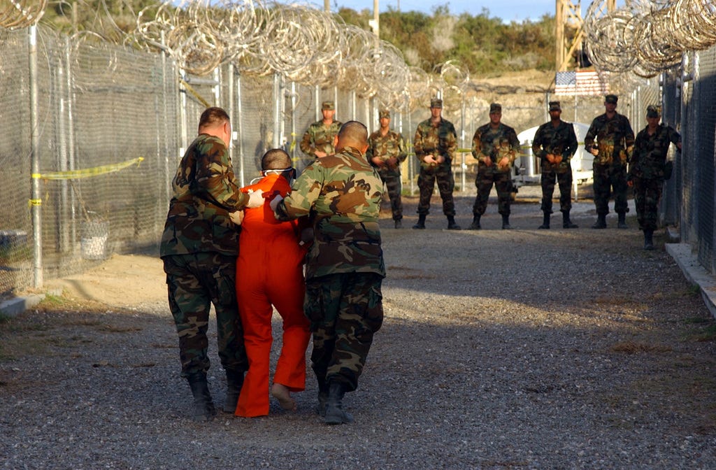 Two US Army (USA) Military Police (MP) escort a detainee, dressed in his  new orange jumpsuit to a cell at Camp X-Ray, Guantanamo Bay Navy Base,  Cuba. Camp X-Ray is the holding