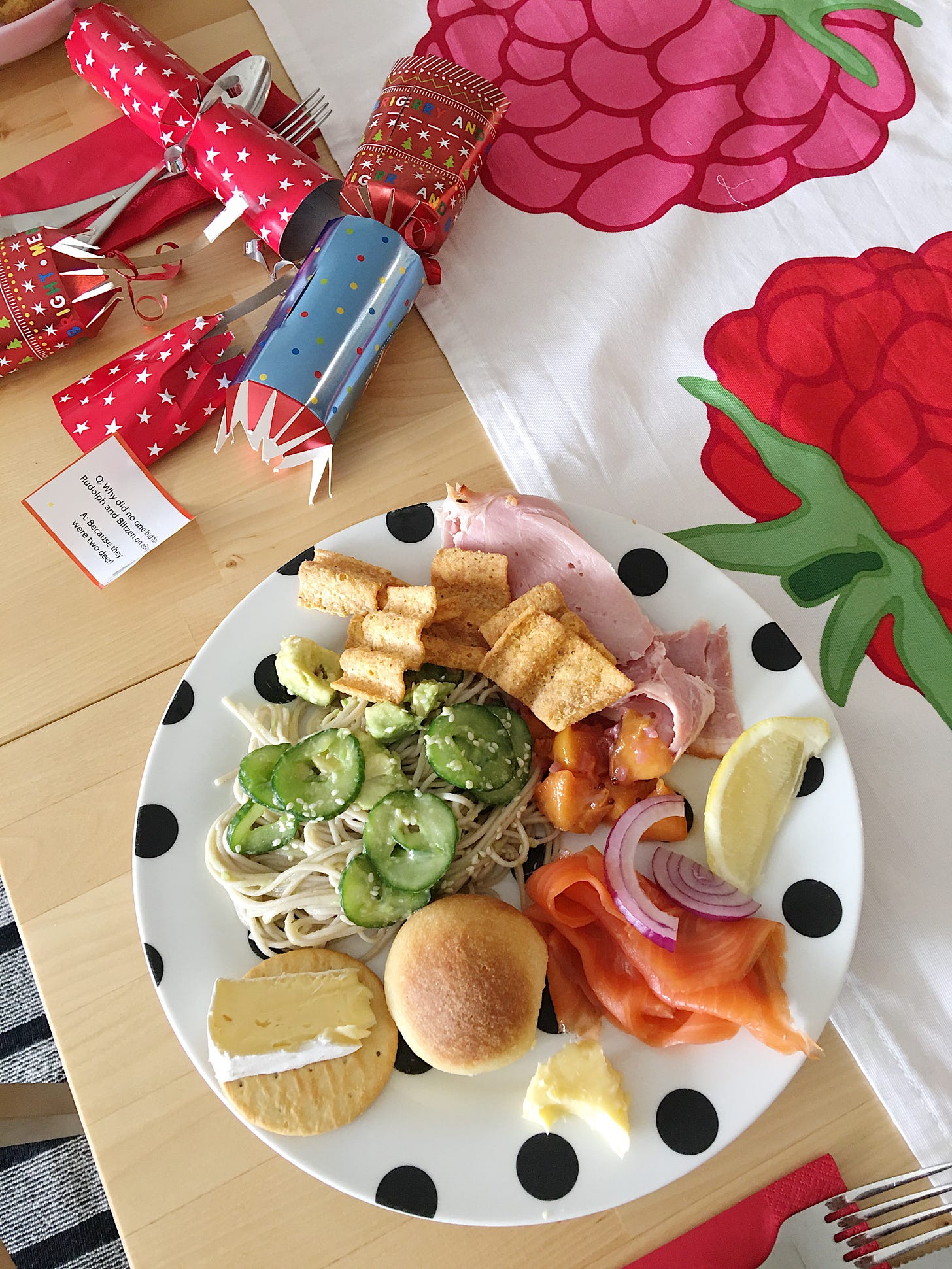 Christmas plate with sliced ham, smoked salmon, grain waves, cheese and crackers and a serving of soba noodle salad. An open Christmas cracker nearby.