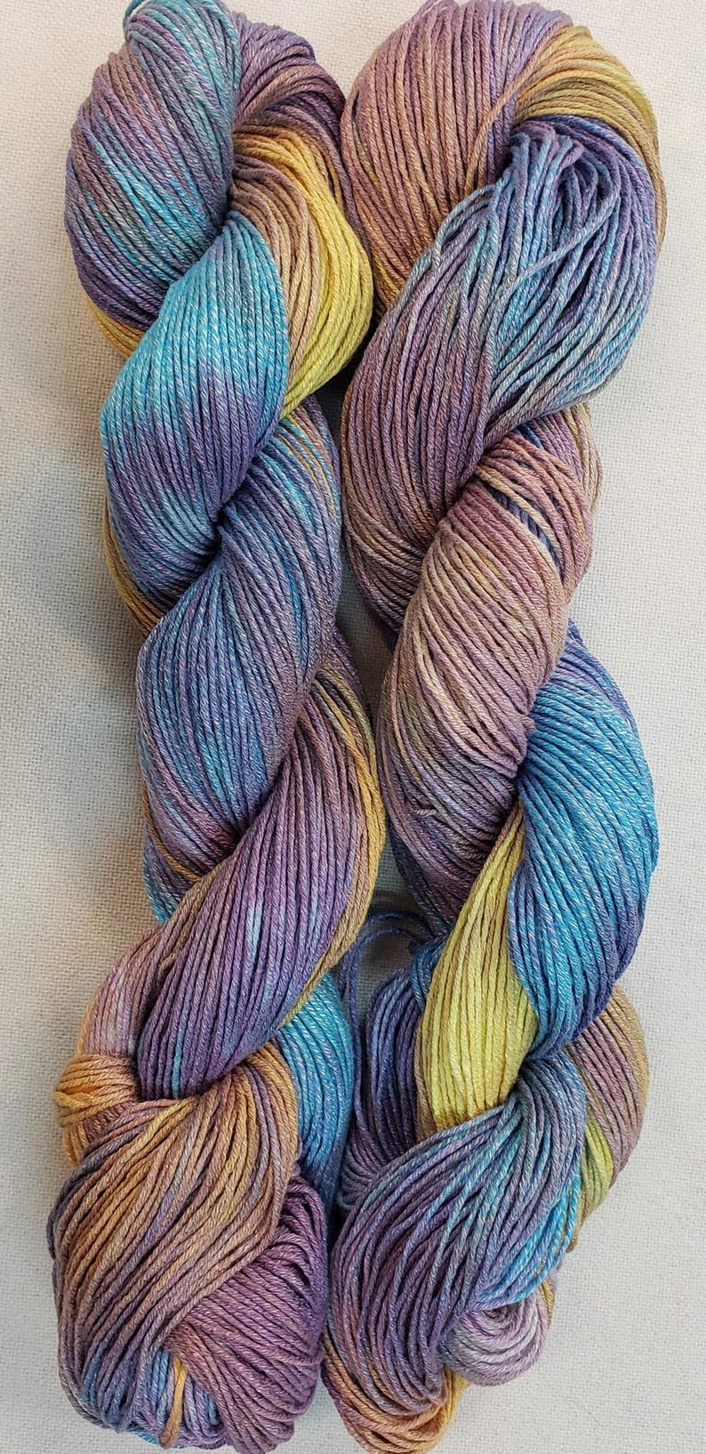 May include: Two skeins of yarn in a multi-colored pattern of purple, blue, yellow, and orange. The yarn is twisted together and appears to be soft and textured.