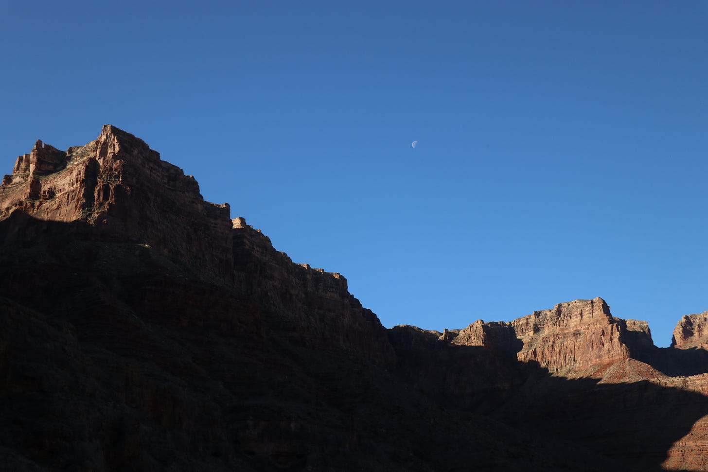 A half moon sits in a dimming evening sky above the rim of the Grand Canyon, half in shadow and half in light