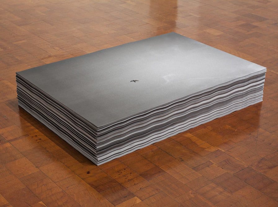 An installation view of Felix Gonzalez-Torres, "Untitled", 1992/1993, a thick stack of paper posters with the black and white photographic image of a small bird in flight, silhouetted against a wide grey sky. The stack is sitting on a rich wooden floor.