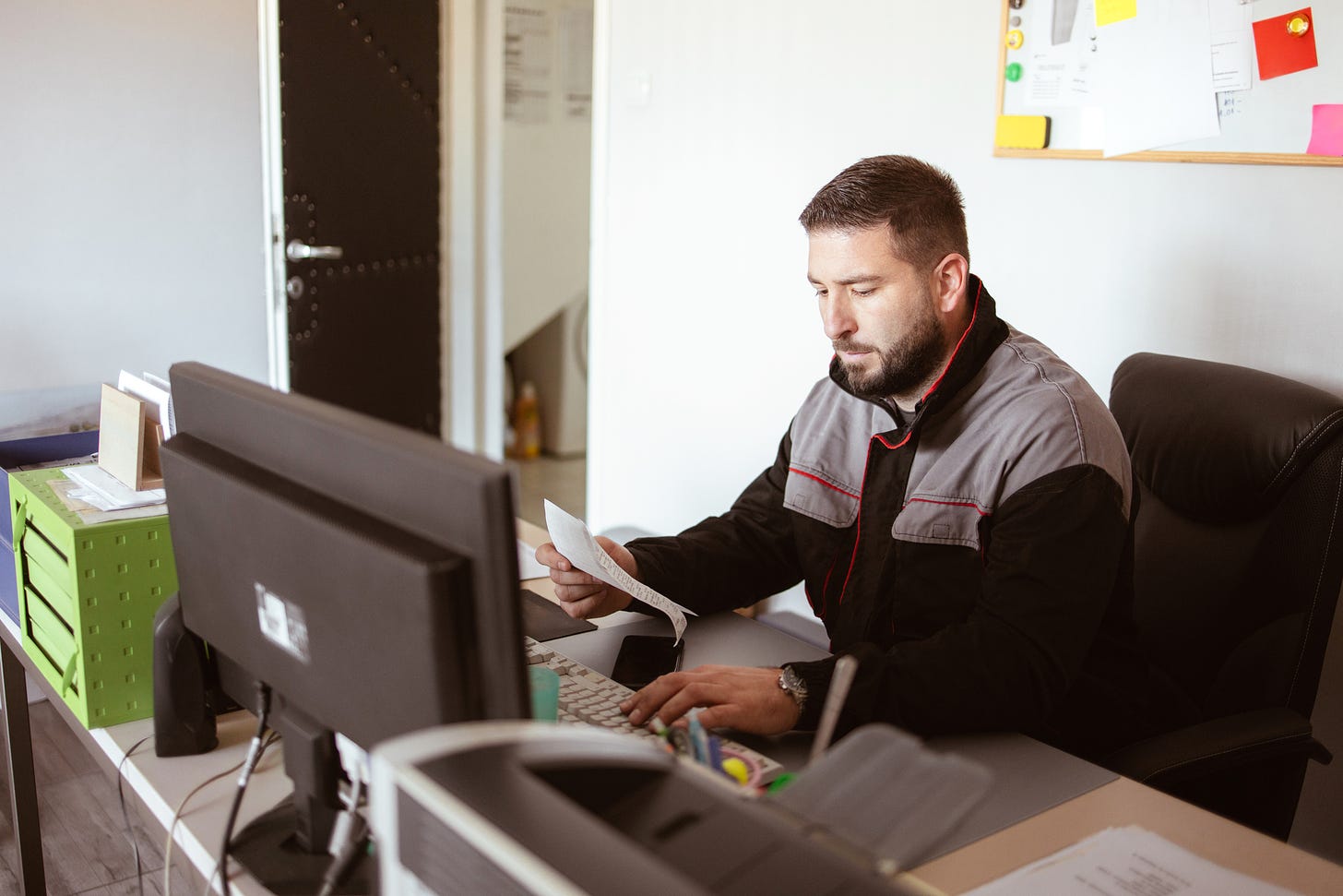 The car service technician writes notes while working on the computer in the offices and checks the invoice issued by the client.