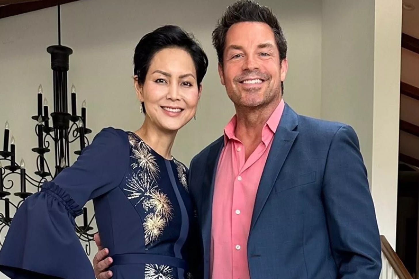 Brennan Elliott poses wearing a blue suit and pink shirt with his wife Camilla Row, wearing a dark blue dress with gold stars