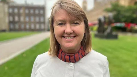 BBC Sarah Gibson smiling at the camera in front of the Houses of Parliament in Westminster
