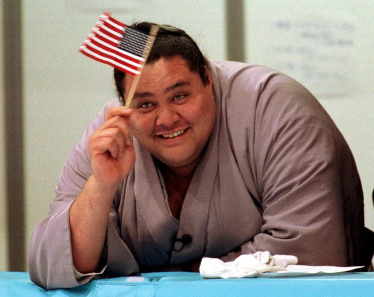 FILE PHOTO: US born Grand Champion sumo wrestler Akebono waves an American flag as he watches US skaters at the White Ring Stadium in Nagano February 5,1998 ahead of the XVIII Winter Olympics./File Photo
