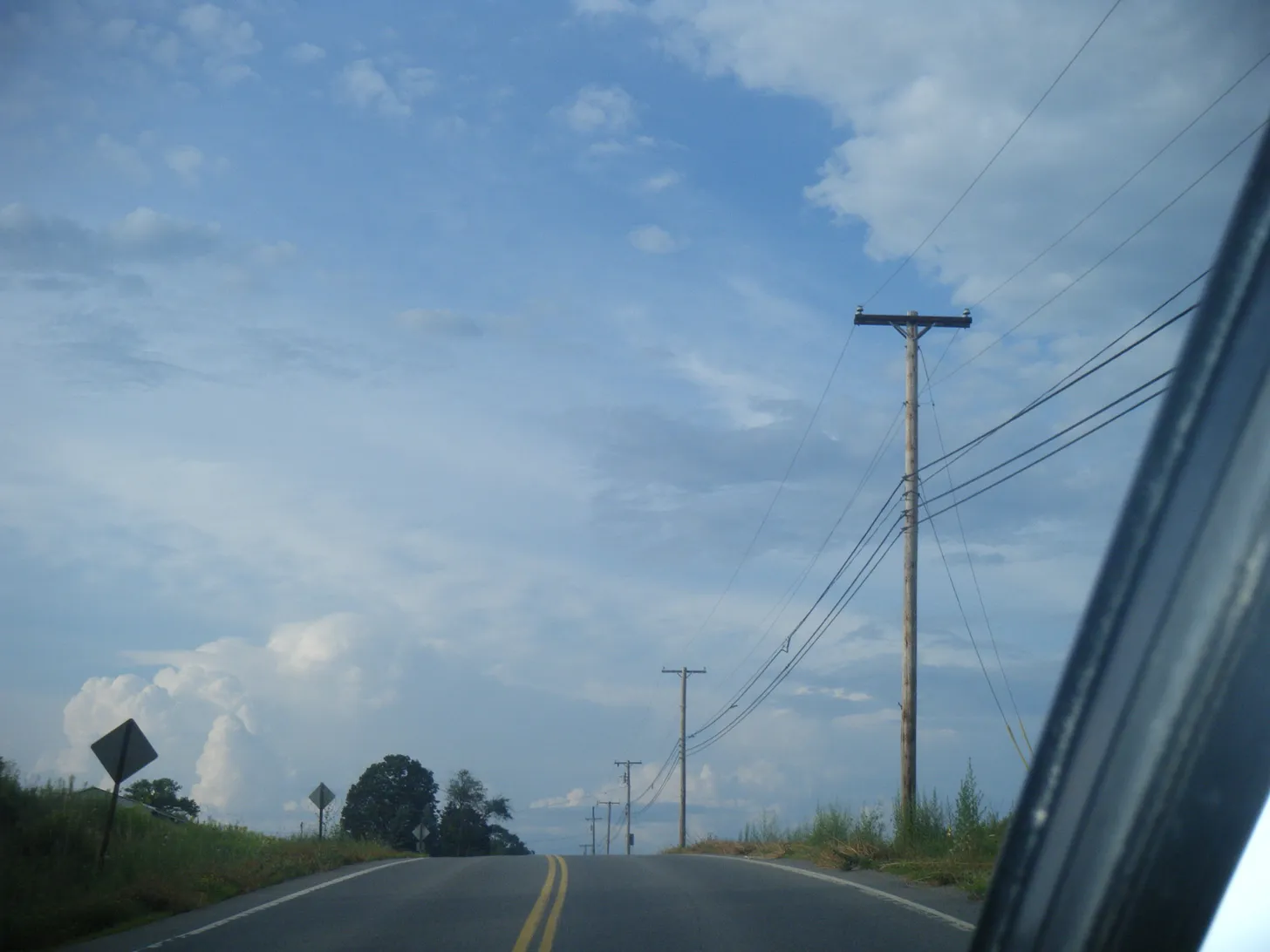 A photograph taken from inside a car of a road. There are power lines strung up to the side, and the main focus is the big blue sky ahead, filled with clouds.