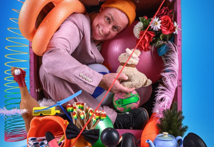 A man in pink suit and orange hat crammed into a box with an odd selection of objects including toys and instruments