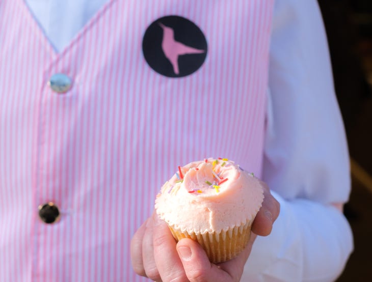 Someone in a Hummingbird Bakery pink striped uniform holding a pink cupcake