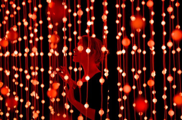 A woman standing among vertical hanging cords adorned with glowing red light bulbs