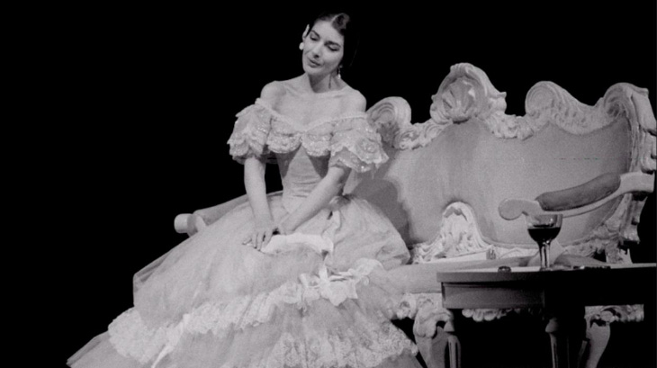 A black and white photograph of Maria Callas, sitting down in a large dress