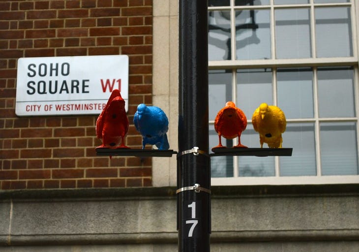 A sculpture of four colourful pigeons sitting on a lampost, in front of a Soho Square sign