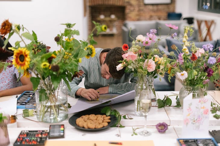 A man sitting at a table with his head down, drawing, surrounding by floral arrangements in vases