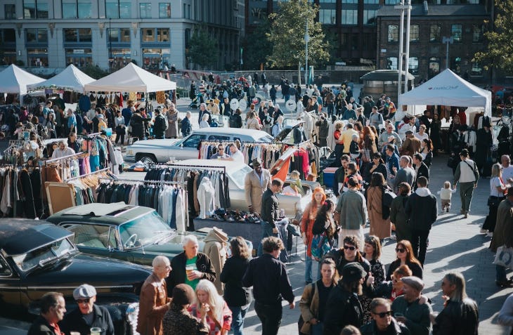 People browsing rails of vintage clothing among parked classic cars