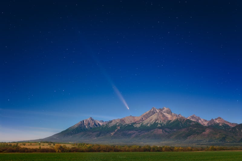 Brightest comet of 2025: Long-tailed comet in a deep cobalt blue sky with a few stars, over some picturesque jagged brown mountains.