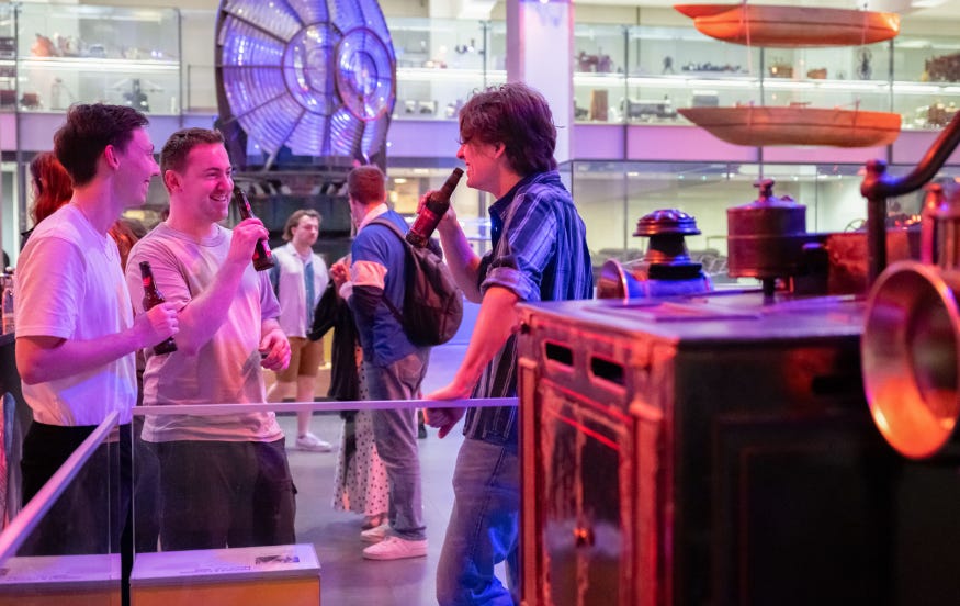 Three men standing chatting and drinking alongside a museum display