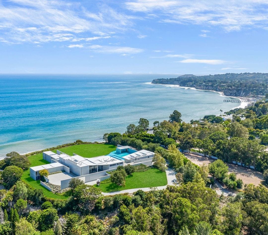 A birds-eye view of a white, sprawling mansion with a pool. It is on a green, tree-filled hillside overlooking the ocean.