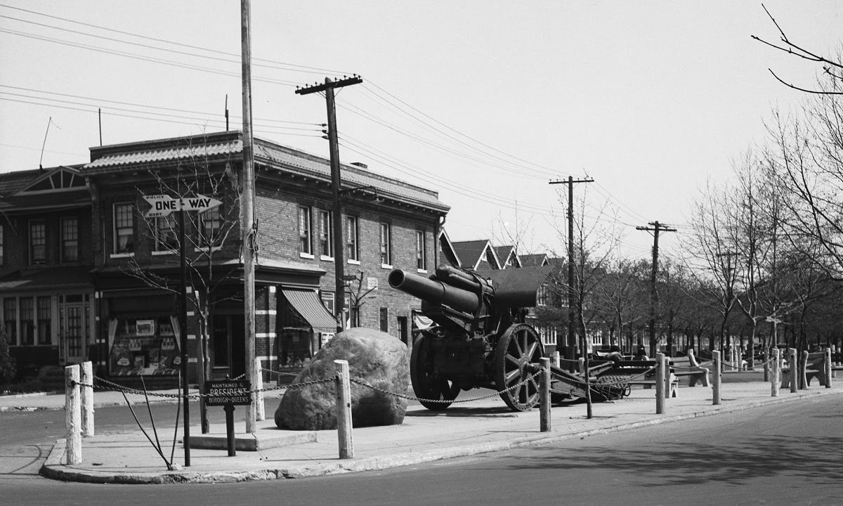Image is photograph of Whiting Square with Cannon, rock, flagpole and a corner store and a row of houses in the background.