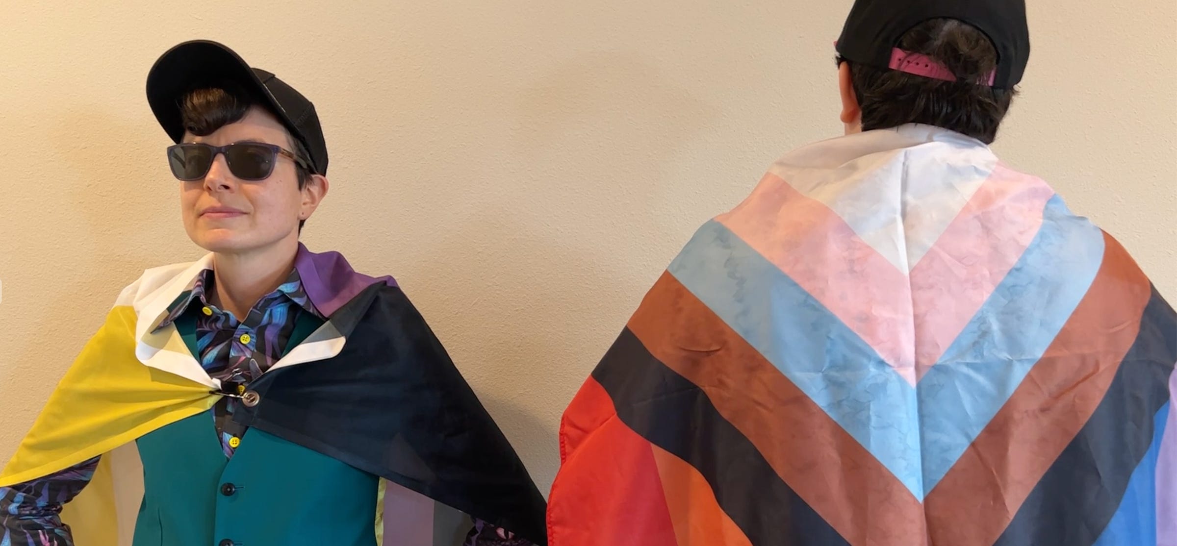 A photo of two white genderqueer people standing in front of a white wall, from the waist up. The person on the left is facing the camera, the other is facing away. They are both wearing pride flags as capes, a nonbinary flag on the left and the progress pride flag on the right. 