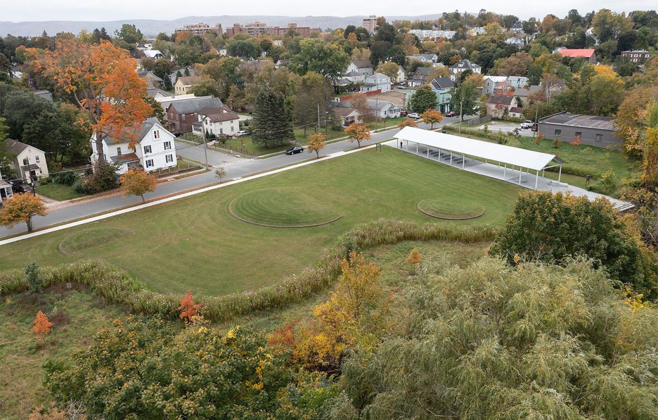 A drone-eye view of a park with several circular mounds in the grass and a white-roofed open pavilion along one edge